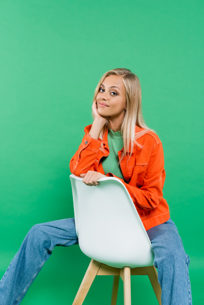 positive blonde woman in orange jacket looking at camera while sitting on white chair isolated on green - Zdjęcie, obraz