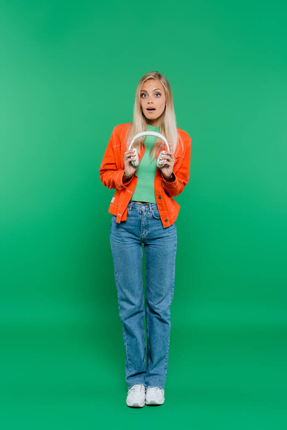 full length view of amazed woman in orange jacket and jeans holding headphones on green - Foto, Imagen