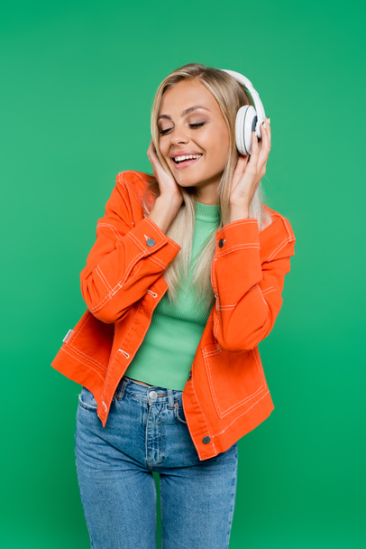 cheerful woman in orange jacket and headphones listening music with closed eyes isolated on green - Zdjęcie, obraz