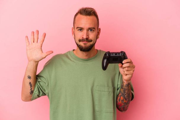 Jovem caucasiano com tatuagens segurando controlador de jogo isolado no fundo rosa sorrindo alegre mostrando número cinco com os dedos. - Foto, Imagem