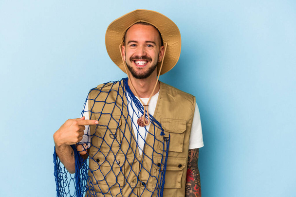 Joven pescador caucásico con tatuajes sosteniendo red aislada sobre fondo azul persona señalando a mano a un espacio de copia de la camisa, orgulloso y confiado - Foto, Imagen