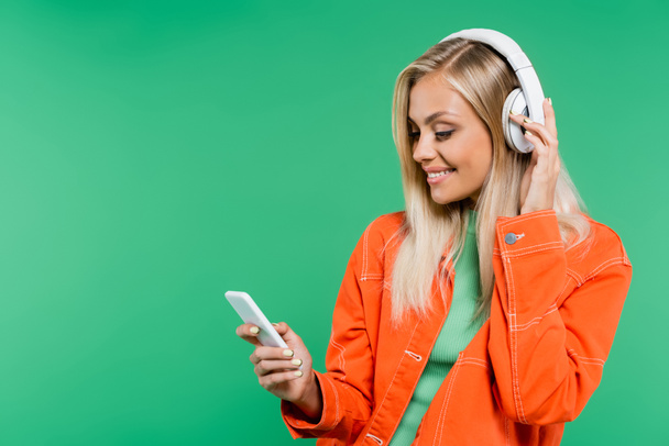 smiling blonde woman looking at mobile phone while listening music in headphones isolated on green - Foto, immagini