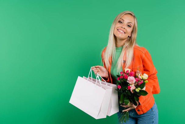vidám nő bevásárló táskák és csokor mosolygó kamera elszigetelt zöld - Fotó, kép