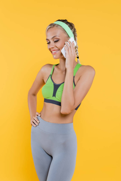 sportive woman in green sports bra and grey leggings standing with hand on hip and talking on smartphone isolated on yellow - Foto, Imagem