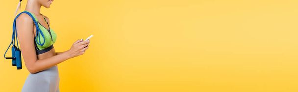 καλλιεργημένη άποψη του χαμογελαστού αθλητικογράφου με σχοινί άλμα στον ώμο χρησιμοποιώντας κινητό τηλέφωνο απομονώνονται σε κίτρινο, banner - Φωτογραφία, εικόνα