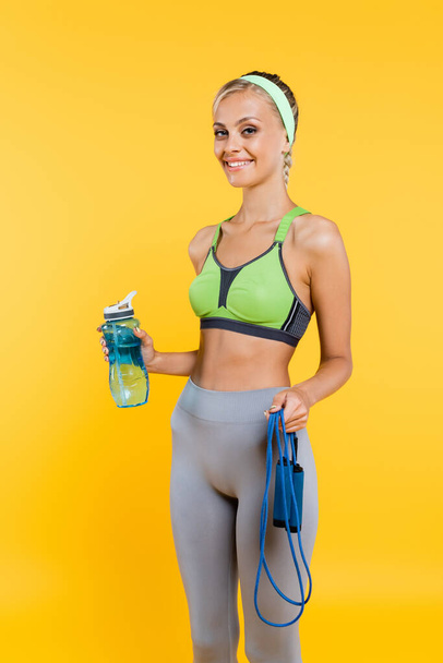 cheerful woman in sportswear holding skipping rope and sports bottle isolated on yellow - Foto, Bild