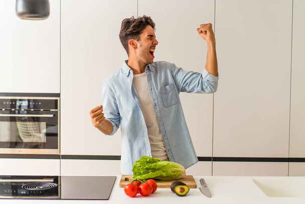 Young mixed race man preparing a salad for lunch raising fist after a victory, winner concept. - Photo, Image