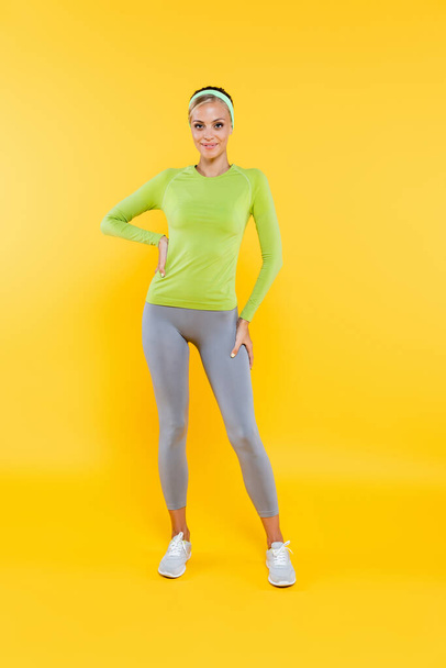 full length view of pretty woman in sports leggings and long sleeve t-shirt standing with hand on hip on yellow - Foto, immagini