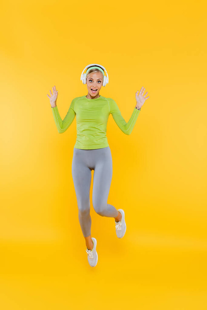 full length view of excited and sportive woman in headphones looking at camera while jumping on yellow - Photo, Image