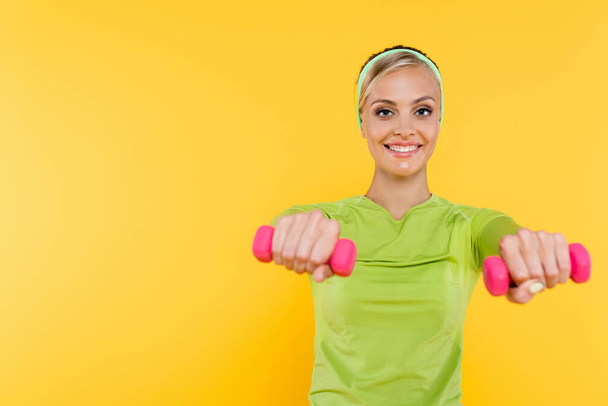 slim woman in green long sleeve shirt smiling while working out with dumbbells isolated on yellow - Photo, Image