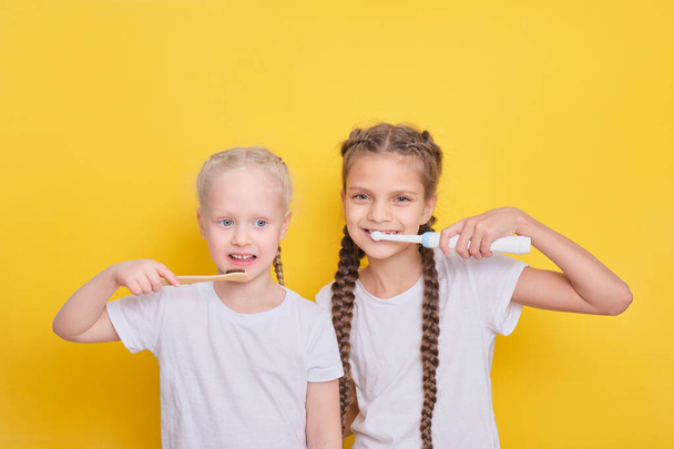 cheerful girls brushing teeth on yellow background, toddler and teen girls, electric or bamboo brush concept, toothbrushes for kids - Foto, Imagen