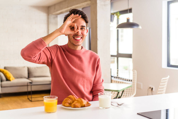 młody mieszane wyścig człowiek mając śniadanie w kuchni w godzinach porannych podekscytowany utrzymanie ok gest na oku. - Zdjęcie, obraz