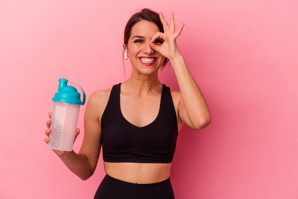 Nuori valkoihoinen nainen juo proteiinipirtelö eristetty vaaleanpunainen tausta innoissaan pitää ok ele silmään. - Valokuva, kuva