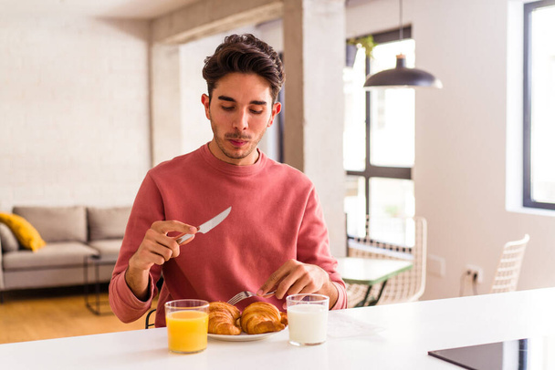 Young mixed race man eating croissant in a kitchen on the morning - Photo, Image