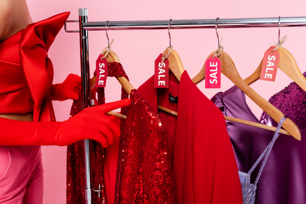 vista cortada de mulher em luvas vermelhas apontando para roupas elegantes com etiquetas de venda isoladas em rosa - Foto, Imagem