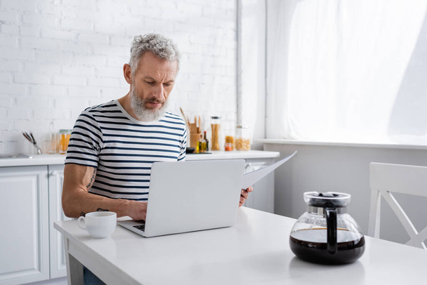 Hombre maduro sosteniendo papeles y utilizando el ordenador portátil cerca de café en casa  - Foto, imagen