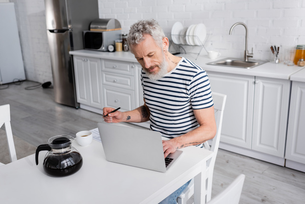 Зрелый мужчина держит ручку и использует ноутбук возле кофе и горшок на кухне  - Фото, изображение