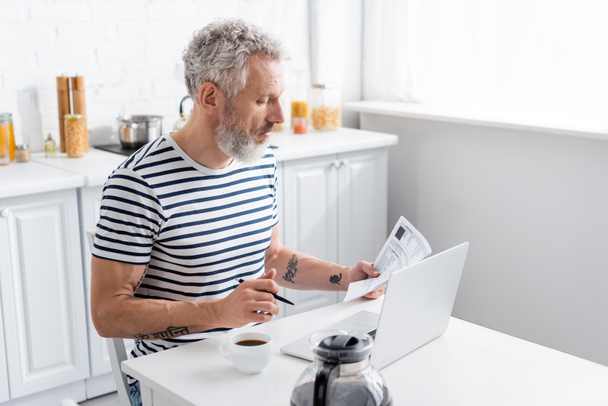 Vue latérale d'un homme d'âge moyen tenant du papier avec des factures et un stylo près d'un ordinateur portable et du café à la maison  - Photo, image