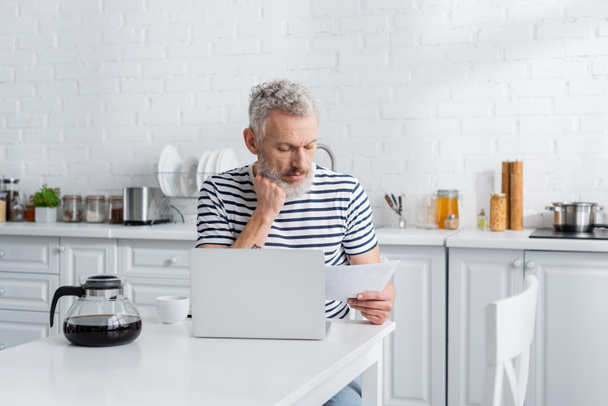 Hombre de mediana edad mirando el documento cerca de café y portátil en la cocina  - Foto, imagen