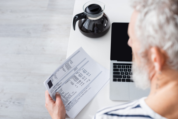 Blurred mature man holding papers with bills near laptop and coffee pot  - Фото, зображення