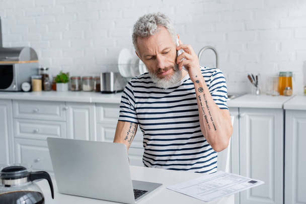Tattooed mature man talking on mobile phone near bills and laptop in kitchen  - Photo, Image