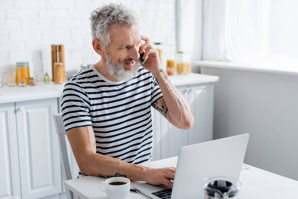 Positive mature freelancer talking on smartphone and using laptop near coffee in kitchen  - Photo, image