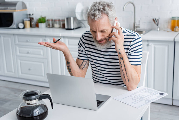 Ώριμος τηλεργάτης μιλάει σε smartphone κοντά σε χαρτιά και laptop στην κουζίνα  - Φωτογραφία, εικόνα
