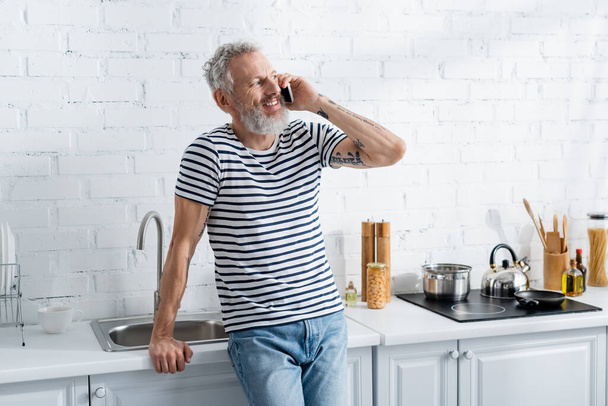 Happy mature man talking on smartphone near worktop in kitchen  - Foto, Imagem