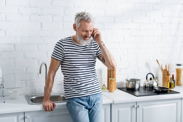 Starší muž mluví na mobilu v kuchyni. Překlad: "Musíte kopat hluboké studny, pokud chcete čistou vodu" - Fotografie, Obrázek