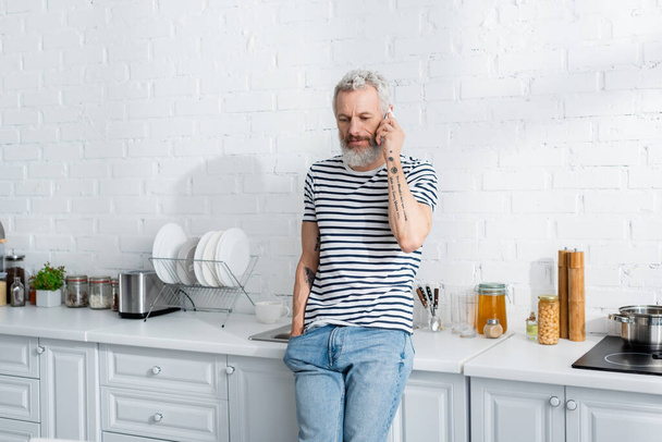 Μεσήλικας μιλάει στο κινητό κοντά στον πάγκο εργασίας στην κουζίνα  - Φωτογραφία, εικόνα