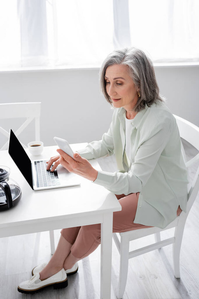 Volwassen vrouw met behulp van laptop en smartphone in de buurt van koffie thuis  - Foto, afbeelding
