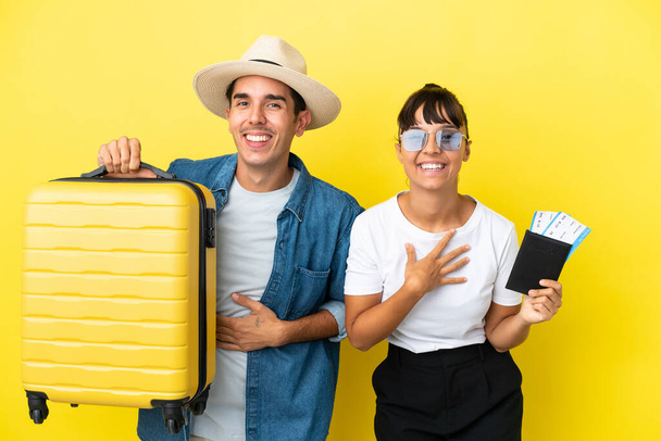Jóvenes amigos viajeros sosteniendo una maleta y pasaporte aislados sobre fondo amarillo sonriendo mucho mientras ponen las manos en el pecho - Foto, Imagen