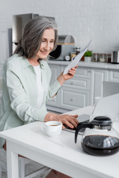 Mujer sonriente sosteniendo facturas y el uso de ordenador portátil cerca de café en la cocina  - Foto, imagen