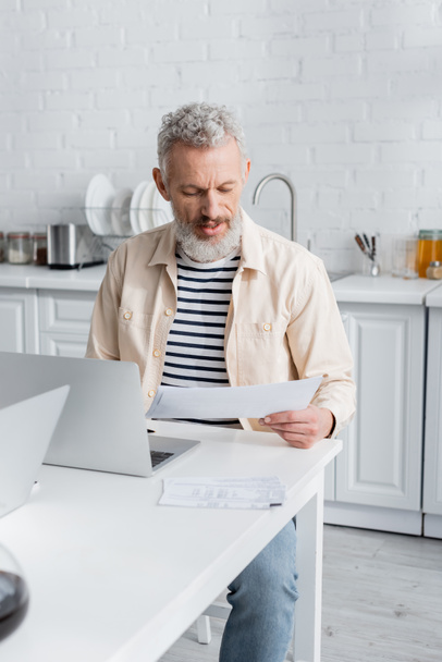 Mature man looking at papers near laptop in kitchen at home  - Foto, afbeelding