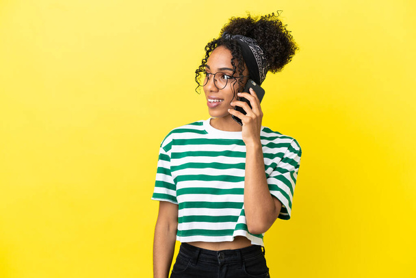 Молодая африканская американка, изолированная на желтом фоне, поддерживает разговор с кем-то на мобильном телефоне - Фото, изображение