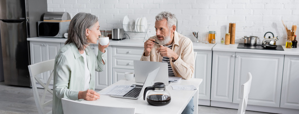 Hombre maduro hablando con su esposa con café cerca de computadoras portátiles y facturas en la cocina, pancarta  - Foto, Imagen