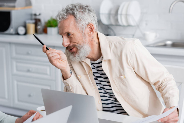 Smiling man holding documents and talking to wife near coffee and laptops in kitchen  - Foto, Bild