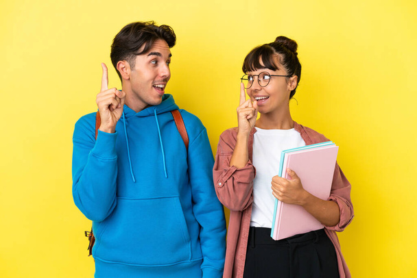 Joven pareja de estudiantes aislados sobre fondo amarillo con la intención de realizar la solución mientras levanta un dedo - Foto, imagen