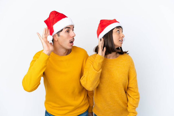 Nuori sekarotuinen pari juhlii joulua eristyksissä valkoisella taustalla kuunnellen jotain laittamalla käden korvaan. - Valokuva, kuva