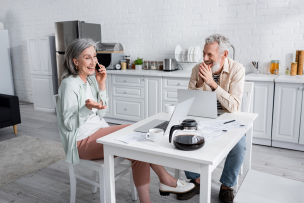 Mujer madura hablando en smartphone cerca de marido alegre, ordenadores portátiles y facturas en la cocina  - Foto, Imagen