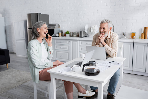 Positive reife Frau spricht auf Smartphone und schaut Ehemann in der Nähe von Laptops und Rechnungen in der Küche an  - Foto, Bild