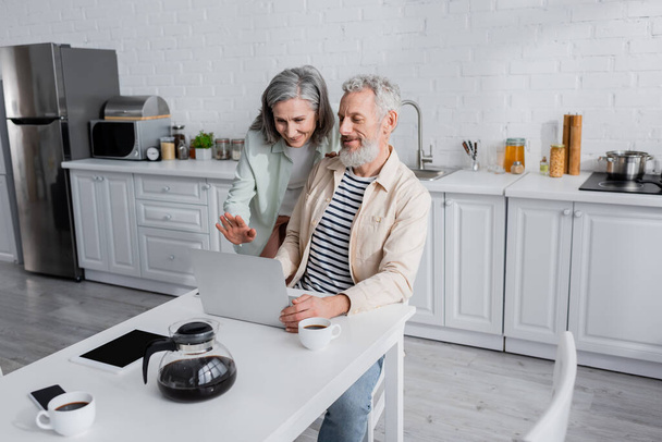 Smiling mature couple having video call on laptop near devices and coffee in kitchen  - Photo, Image