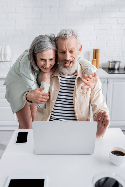 Allegro coppia di mezza età avendo videochiamata sul computer portatile vicino a caffè e dispositivi in cucina  - Foto, immagini