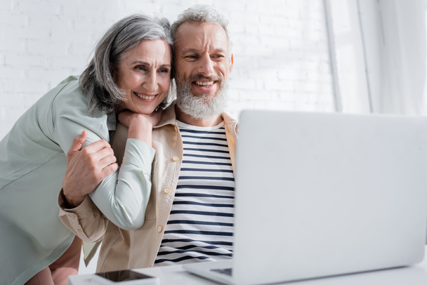Positive mature woman hugging husband and looking at blurred laptop at home  - Photo, Image