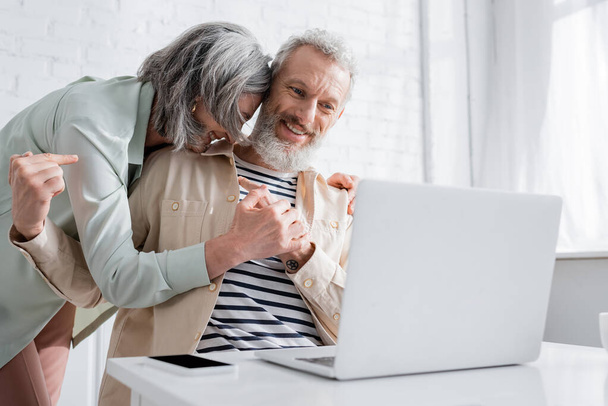 Uśmiechnięty mężczyzna wskazujący żonę podczas rozmowy wideo na laptopie w domu  - Zdjęcie, obraz
