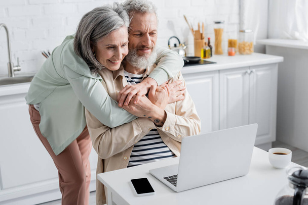 Mature woman holding hand of husband near devices and coffee in kitchen  - Φωτογραφία, εικόνα