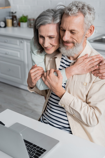 Mature couple hugging near laptop in kitchen at home  - Foto, immagini
