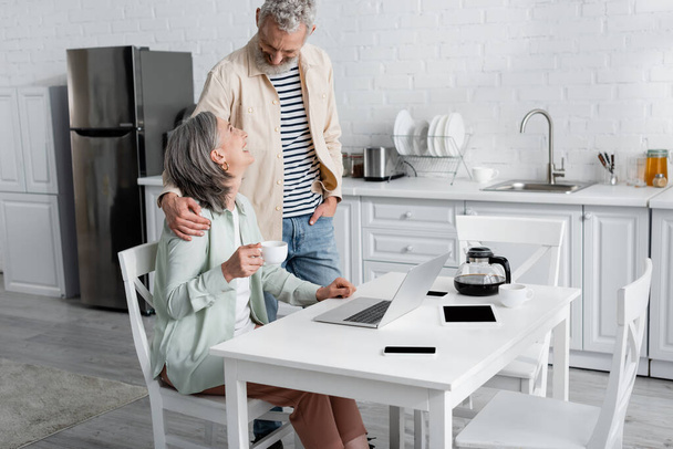 Hombre alegre abrazando esposa con café cerca de gadgets en la cocina  - Foto, Imagen