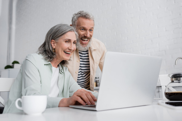 Positive mature couple looking at blurred laptop near coffee pot in kitchen  - Фото, изображение