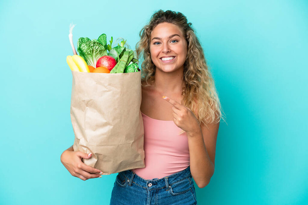 Menina com cabelo encaracolado segurando um saco de compras mercearia isolado no fundo verde apontando para o lado para apresentar um produto - Foto, Imagem